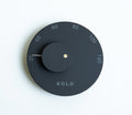 A minimalist round wall clock by Kolo, featuring a matte black face, a central black hub with a small gold center dot, and a thin white hand pointing to 20. The outer ring is marked with white numbers from 20 to 120.