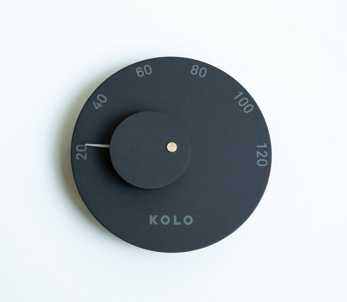 A minimalist round wall clock by Kolo, featuring a matte black face, a central black hub with a small gold center dot, and a thin white hand pointing to 20. The outer ring is marked with white numbers from 20 to 120.